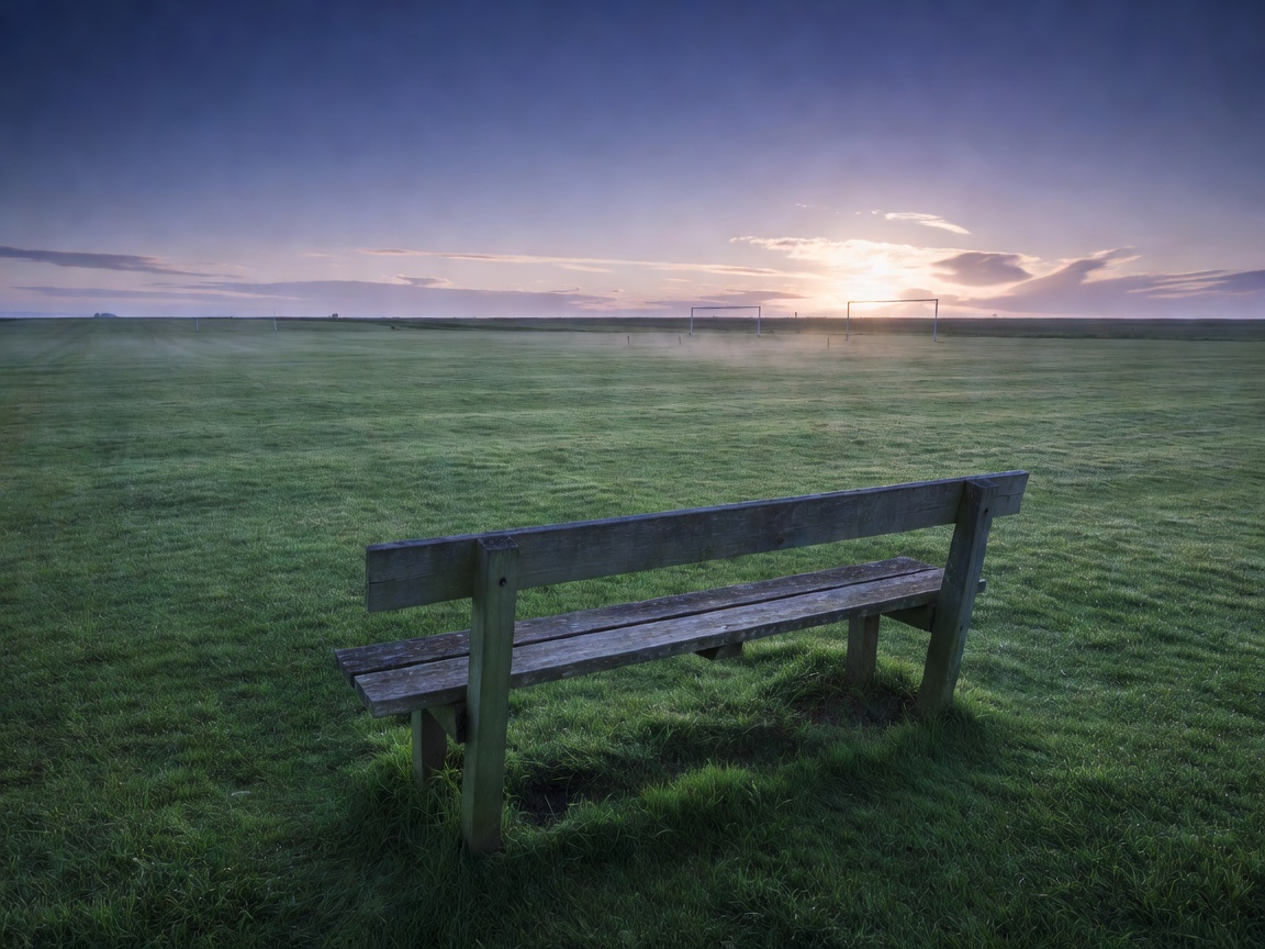 A lone wooden bench beside an empty training pitch at dawn